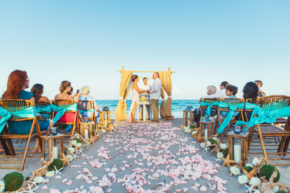 Barefoot bride and groom hold hands during wedding ceremony with pink rose petals, tiffany blue chair sashes, and the blue ocean in the background.