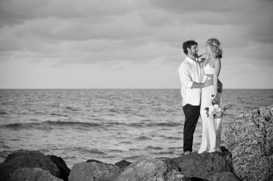 Bride and Groom smile and look into each other's eyes on Miami beach rocks - with ocean in the background