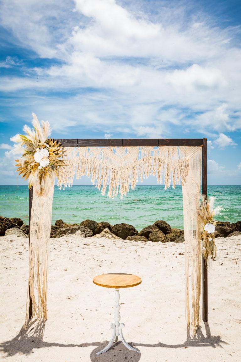 Closeup of Macramé Wedding Arbor with Table, Against turquoise waters, white sand beach, and rocks. Boho Beach Style.