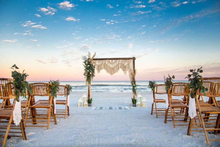 Boho Chic Wedding Package on the Beach - Featuring an Arbor with Lace Accents, Wooden Chairs, and Greenery.
